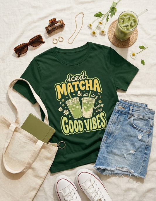 A dark green graphic t-shirt featuring the text 'iced MATCHA & all day GOOD VIBES' with illustrations of two matcha drinks, styled in a flat lay with denim shorts, a canvas tote bag, white sneakers, sunglasses, and a glass of iced matcha on a white fabric background.