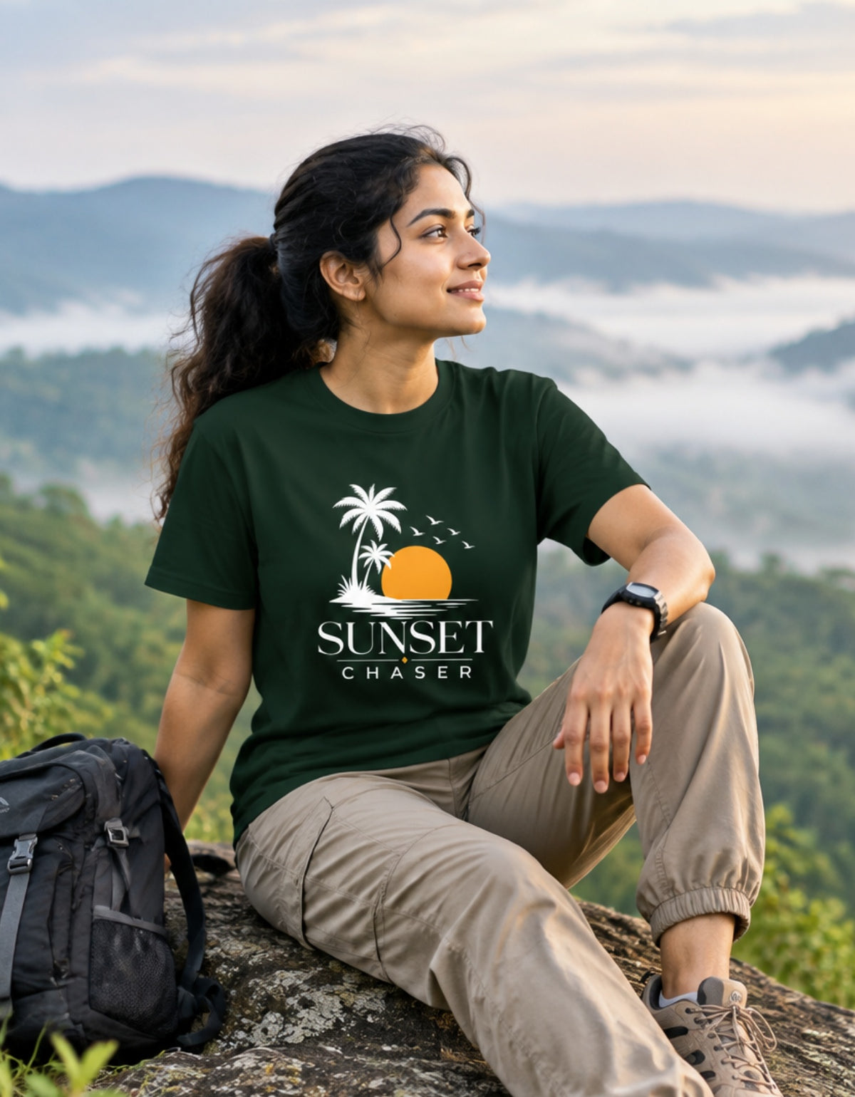 A woman sitting on a rock outdoors wearing a dark green t-shirt with a 'Sunset Chaser' graphic, khaki pants, and a backpack beside her, with misty mountains in the background.