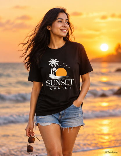A woman stands on a beach at sunset wearing a black graphic T-shirt with a palm tree, sun, and the words 'SUNSET CHASER,' paired with denim shorts and holding sunglasses.