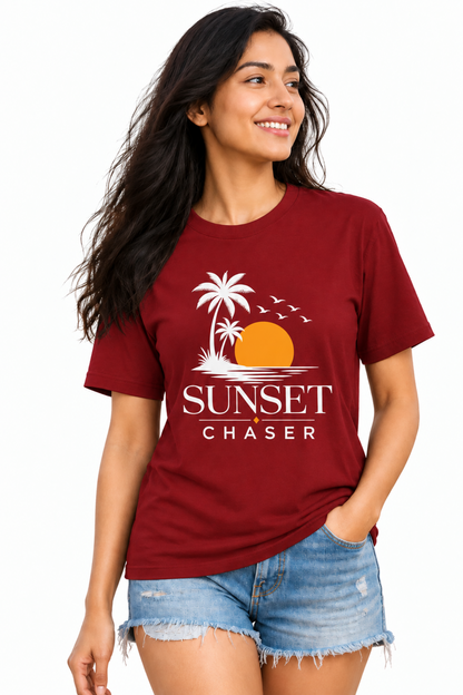 A woman wearing a maroon crewneck t-shirt with a white graphic that reads 'SUNSET CHASER' with palm trees and an orange sun, paired with denim shorts, against a plain white background.