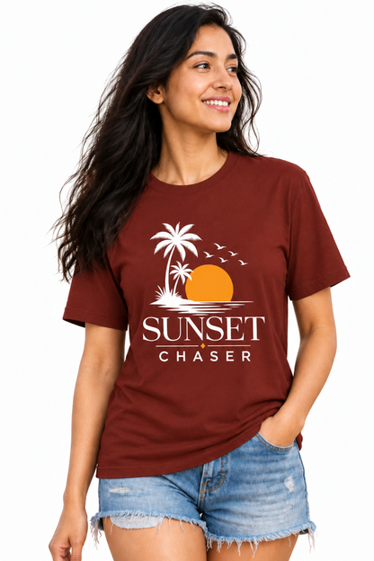 A woman wearing a maroon crewneck t-shirt with a white and orange 'Sunset Chaser' graphic print, paired with denim shorts, against a plain white background.