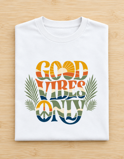 A folded white t-shirt on a light wood background, featuring a colorful retro graphic that reads 'GOOD VIBES ONLY' with palm leaves and a peace sign.