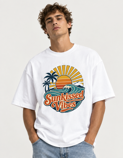 A man wearing a white crewneck t-shirt with a colorful retro graphic that reads 'Sunkissed Vibes' with a sunset, palm trees, and ocean waves, paired with light blue jeans against a light gray background.
