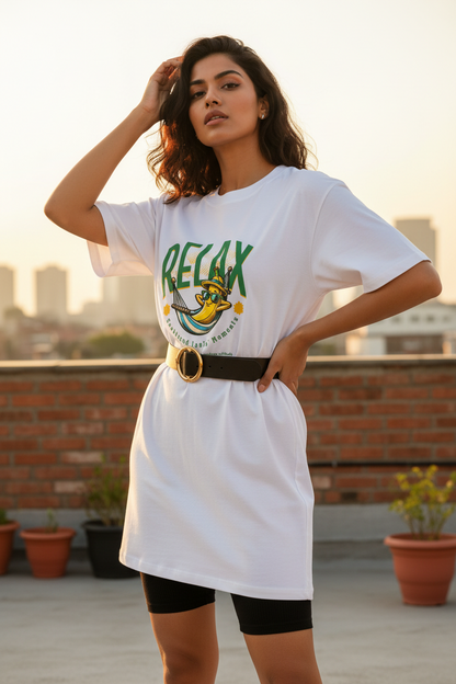 A woman stands on a rooftop wearing a white oversized graphic T-shirt dress with green and yellow 'RELAX' lettering, black fitted shorts, and a black belt with a gold buckle. The background shows a city skyline at sunset with potted plants.