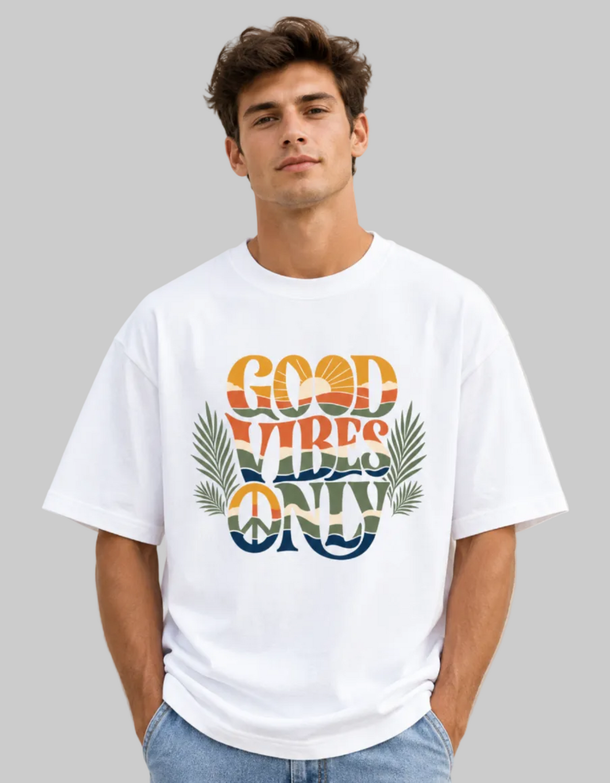 A man wearing a white graphic t-shirt with a retro-style 'GOOD VIBES ONLY' design featuring palm leaves and a peace sign, paired with light blue jeans, set against a solid gray background.