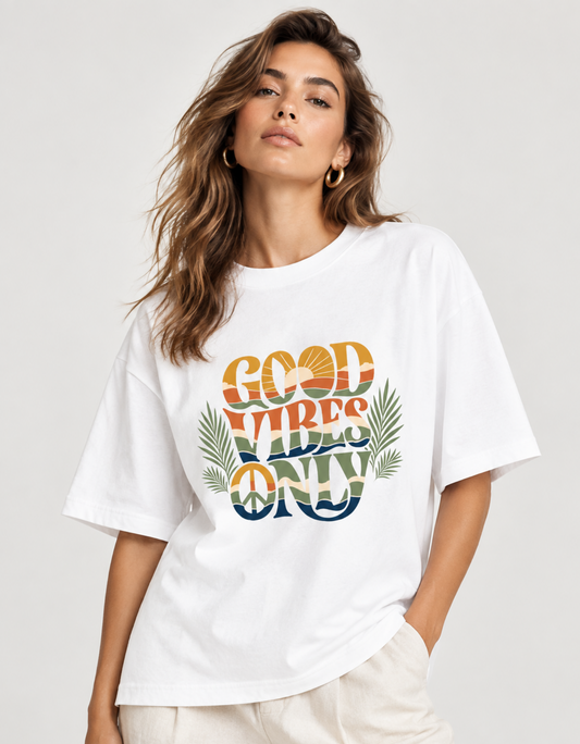 A woman wearing a white oversized graphic T-shirt with the text 'GOOD VIBES ONLY' in a retro sunset gradient and palm leaves, paired with light beige pants against a light gray studio background.
