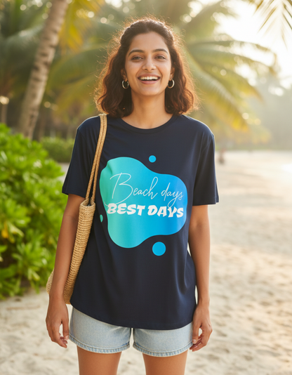 A woman stands on a sandy beach wearing a navy blue graphic t-shirt with a blue and teal splash design that reads 'Beach days BEST DAYS'. She is also wearing light denim shorts and carrying a woven straw shoulder bag, with palm trees and warm sunlight in the background.