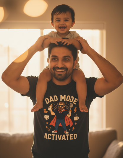 A smiling dad wearing the navy "Dad Mode Activated" superhero graphic T-shirt laughs as his happy toddler sits on his shoulders in a warmly lit living room, illustrating a playful, premium quality gift for new dads, Father's Day, and birthdays.