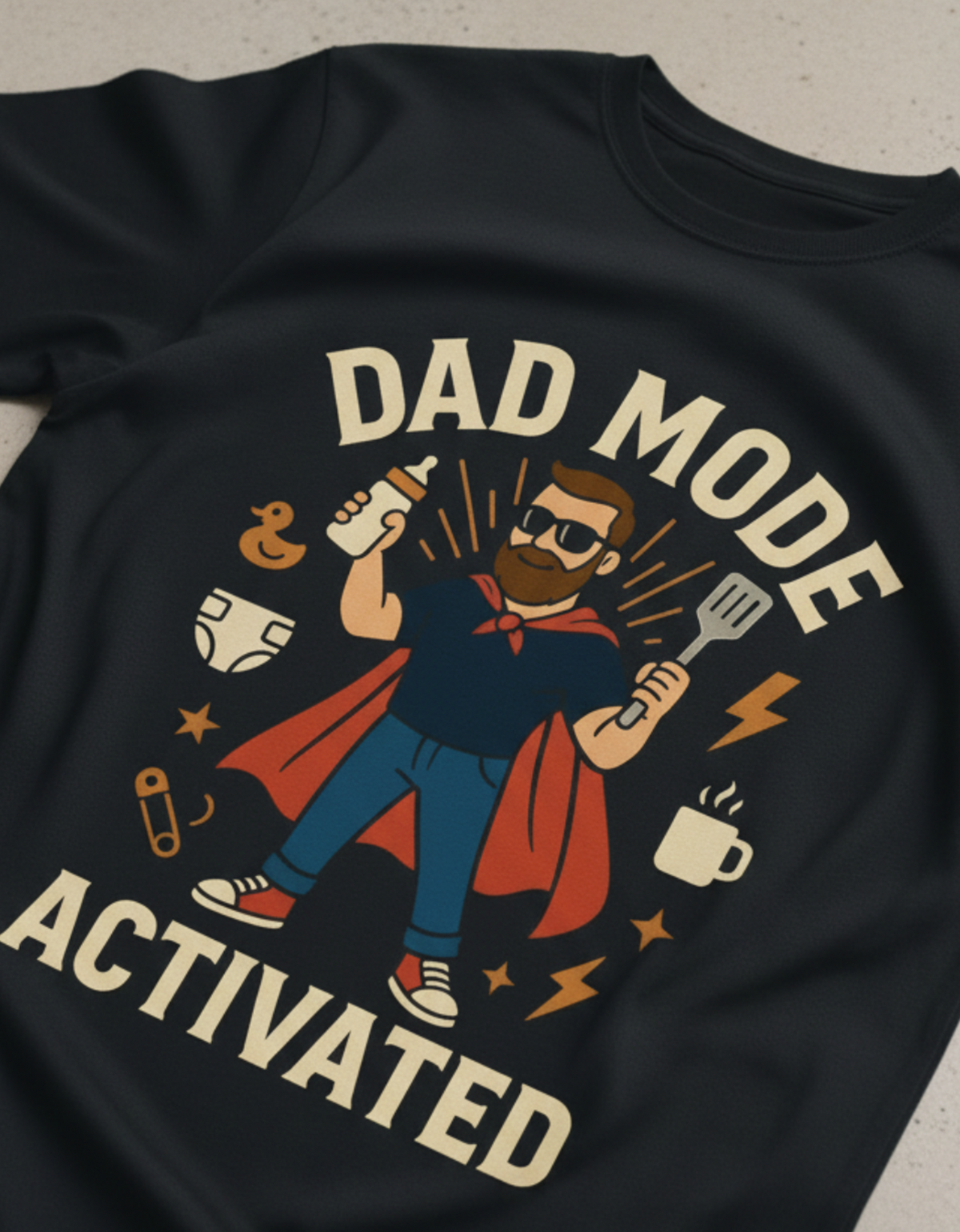A close-up of a dark navy or black t-shirt featuring a colorful graphic print of a superhero dad with the text 'DAD MODE ACTIVATED'. The graphic includes a cartoon dad in a blue shirt and red cape, holding a baby bottle and a spatula, surrounded by icons for a rubber duck, diaper, mug, and safety pins.